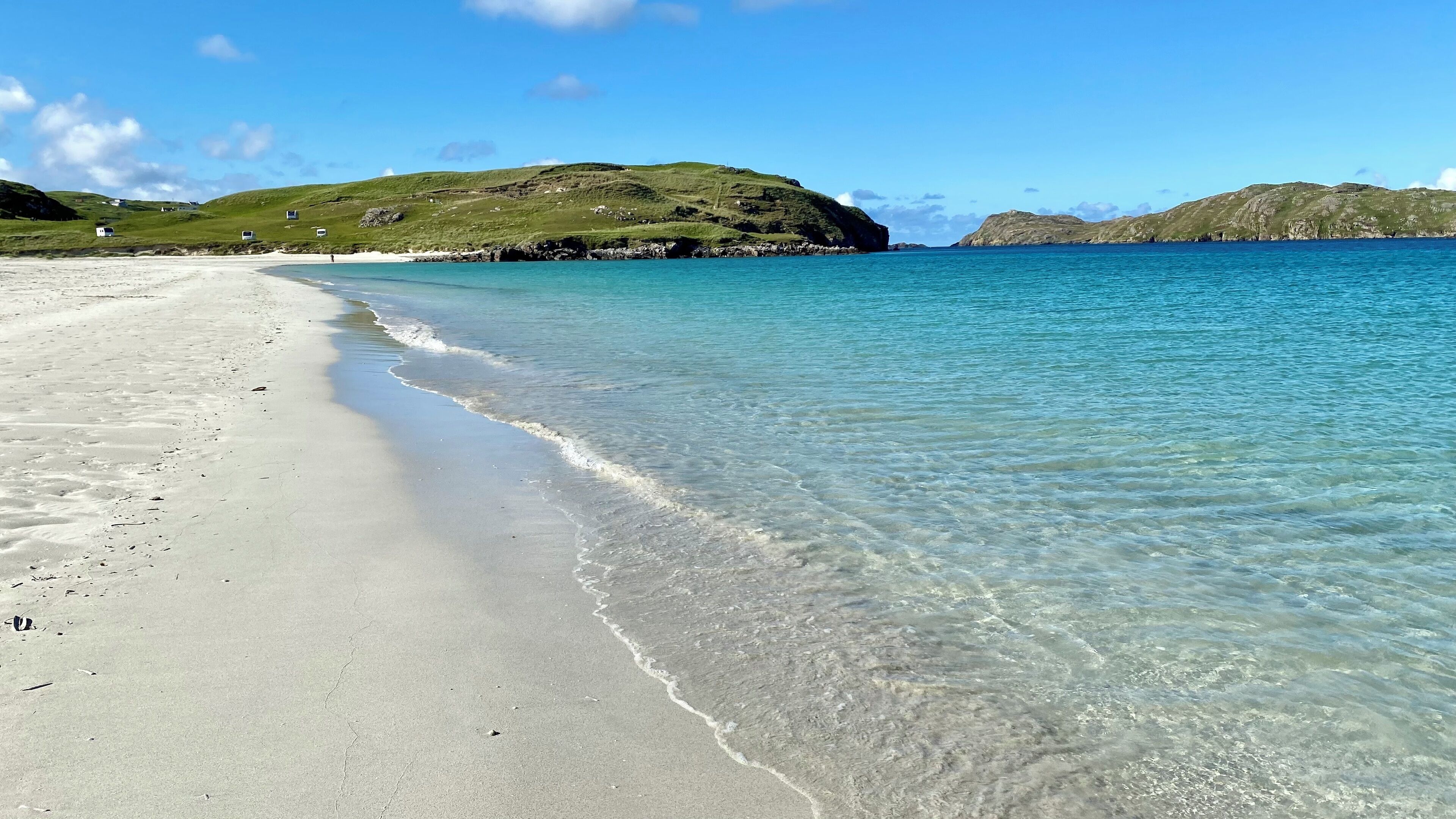 En strand med klart vatten, en sandstrand och kullar i bakgrunden.