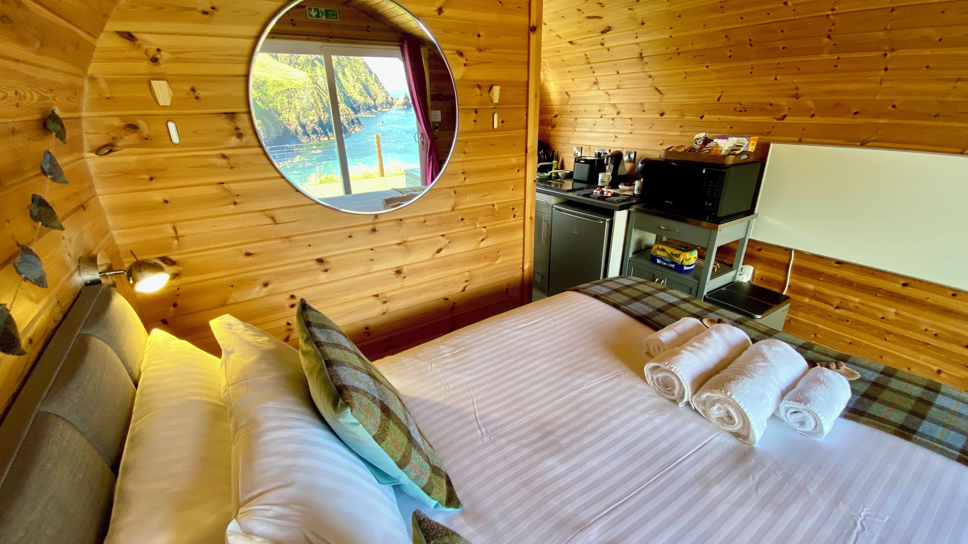 Een houten kamer met een bed, kussens, een spiegel, een kleine kitchenette en uitzicht op zee.