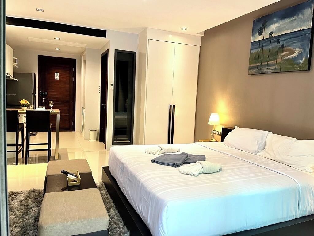 A hotel room with a large bed, a desk with a chair, a small table with a vase, a painting on the wall, and a door leading to another room.