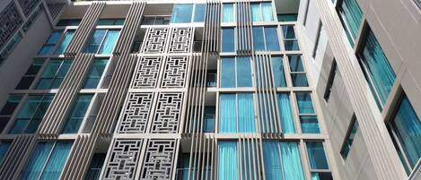 A modern building with a symmetrical facade, featuring a central decorative panel with geometric patterns, multiple windows with blue curtains, and a flat roof with greenery.