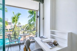 A balcony with a view of a swimming pool and palm trees, a small table with chairs, and a white shelf with a book and a cup.