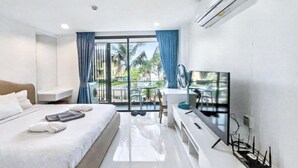 A hotel room with a bed, a TV, a desk, and a balcony with a view of palm trees. - Pixel-modern 1-br Apartment With Pool View in Cape Panwa (Wichit)