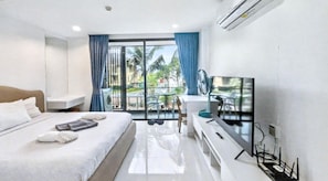 A hotel room with a bed, a TV, a desk, and a balcony with a view of palm trees. - Pixels - Ocean Front Studio With Pool View Cape Panwa (Wichit)