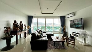 A modern living room with a sofa, coffee table, and a TV mounted on the wall. There is a large window with a view of the city.