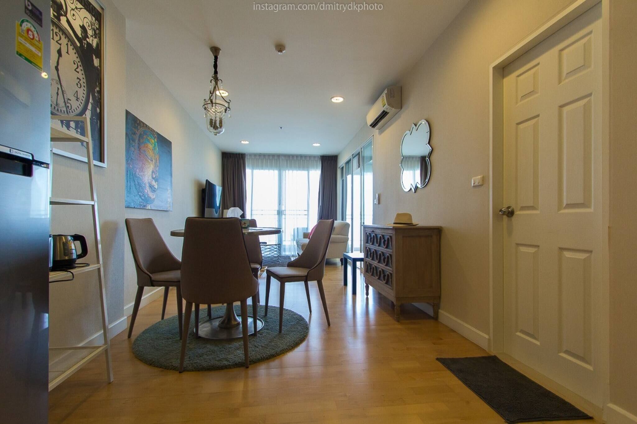 A modern living room with a dining area, wooden flooring, a chandelier, and a large clock on the wall.