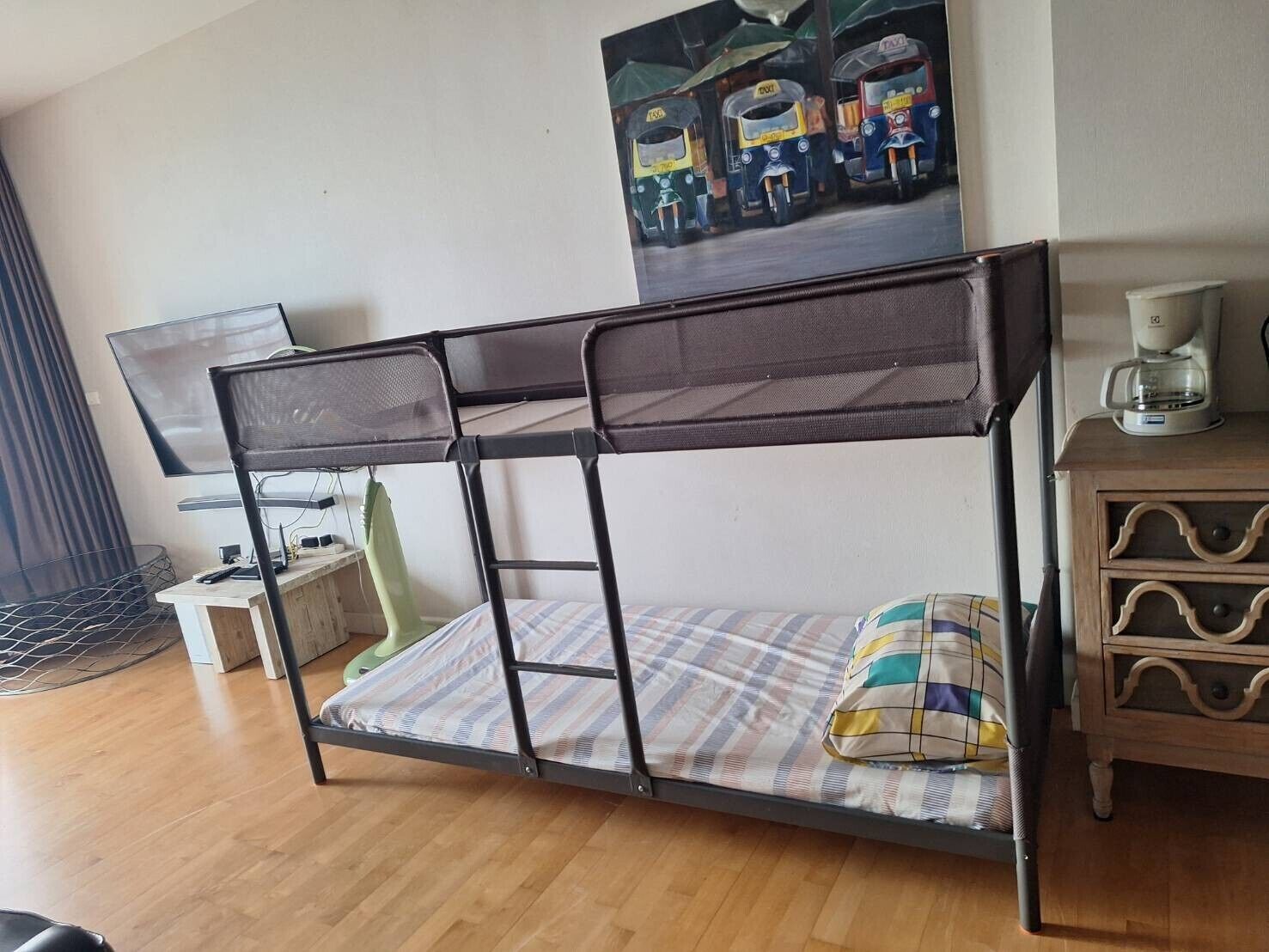Uma cama beliche com colchão e travesseiros, uma mesinha de cabeceira com abajur e um quadro com desenhos de veículos na parede.