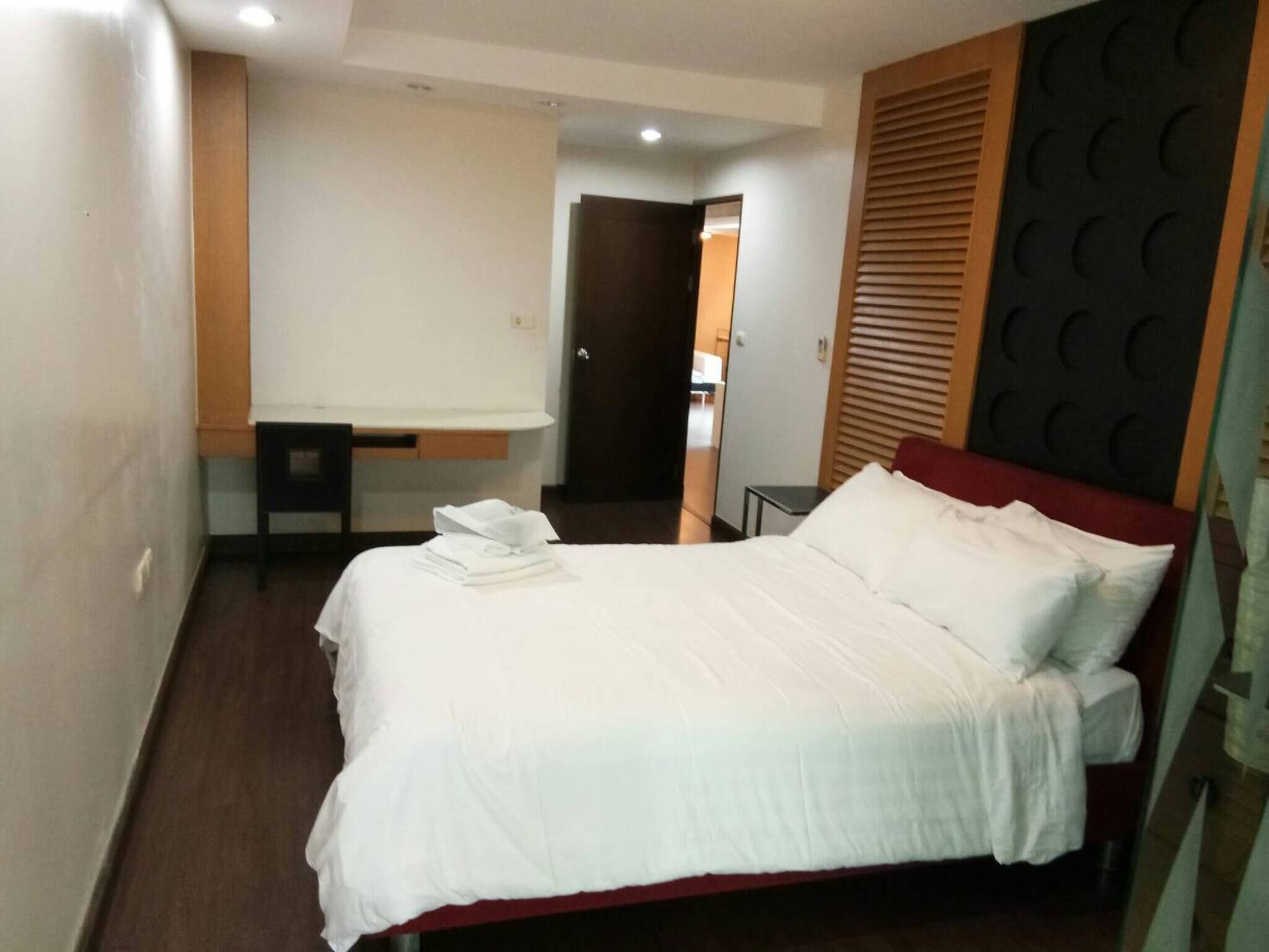 A neatly made bed with white linens and a red headboard, a small bedside table with a white towel, and a wooden panel with circular cutouts on the wall.