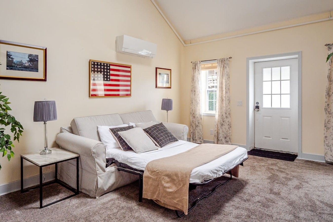 A bedroom with a bed, two bedside tables, a window with curtains, a door, and an air conditioning unit.