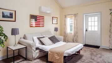 A bedroom with a bed, two bedside tables, a window with curtains, a door, and an air conditioning unit.