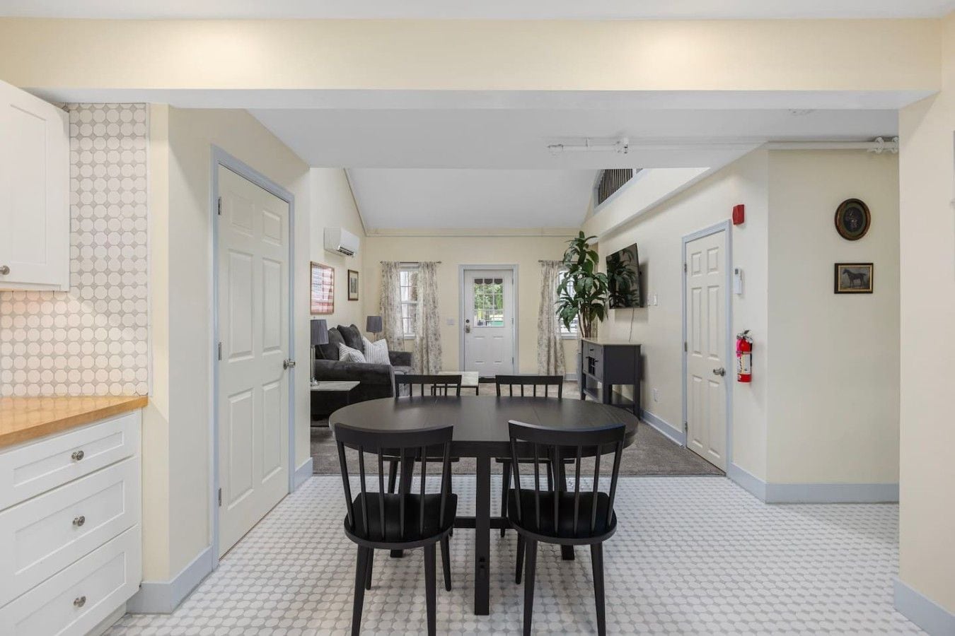 A compact kitchen with a dining area, white tiled flooring, and a view into a living room with a sofa and curtains.