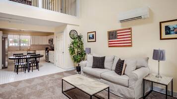 A spacious living area with a sofa, coffee table, and a dining table with chairs. There is a kitchen in the background, a large window, and an American flag on the wall.