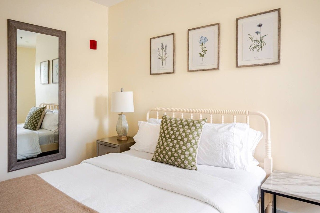 A hotel room with a bed, a nightstand, a lamp, a mirror, and three framed botanical prints on the wall.