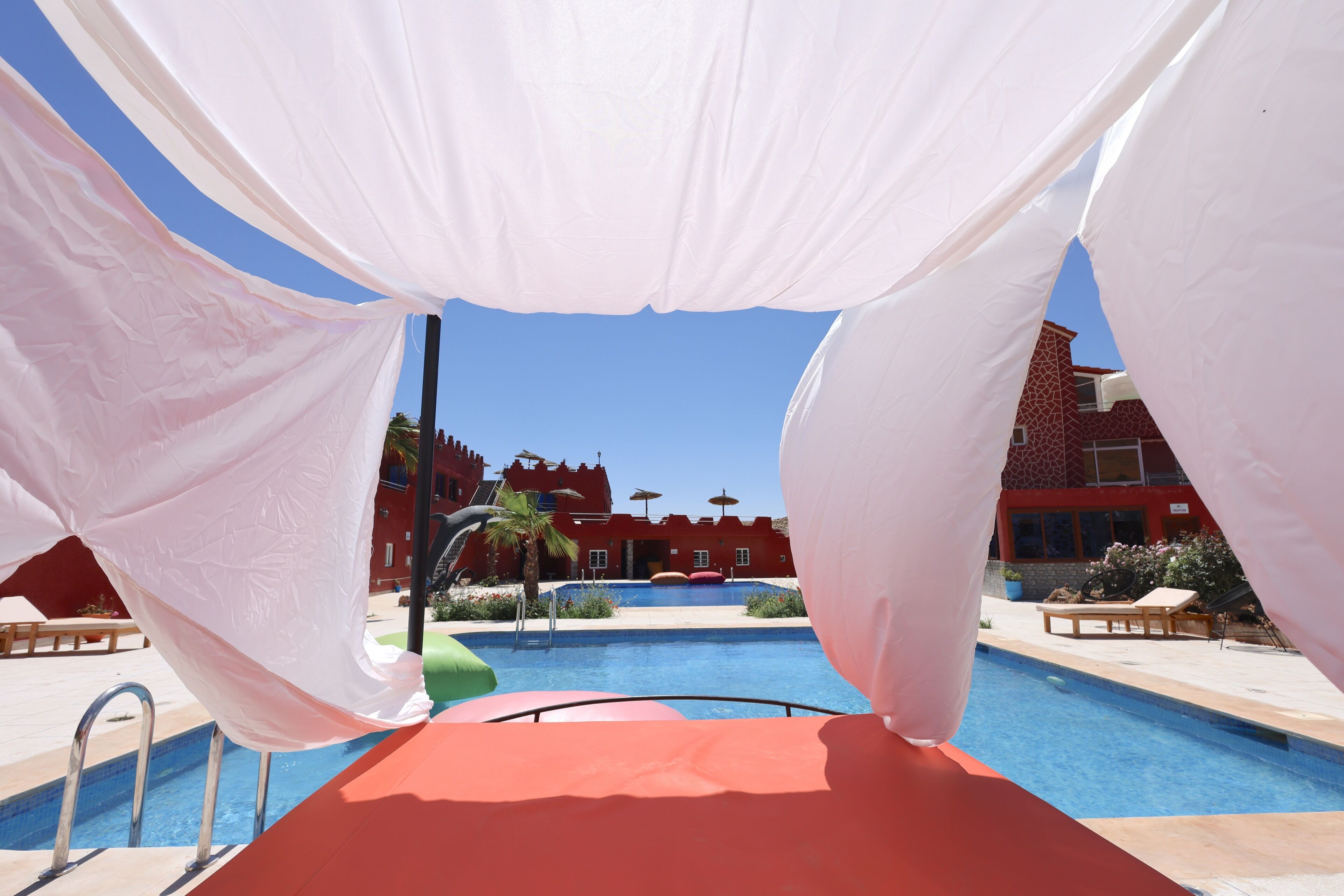 A swimming pool with a red deck, surrounded by white drapes, and a red building in the background.