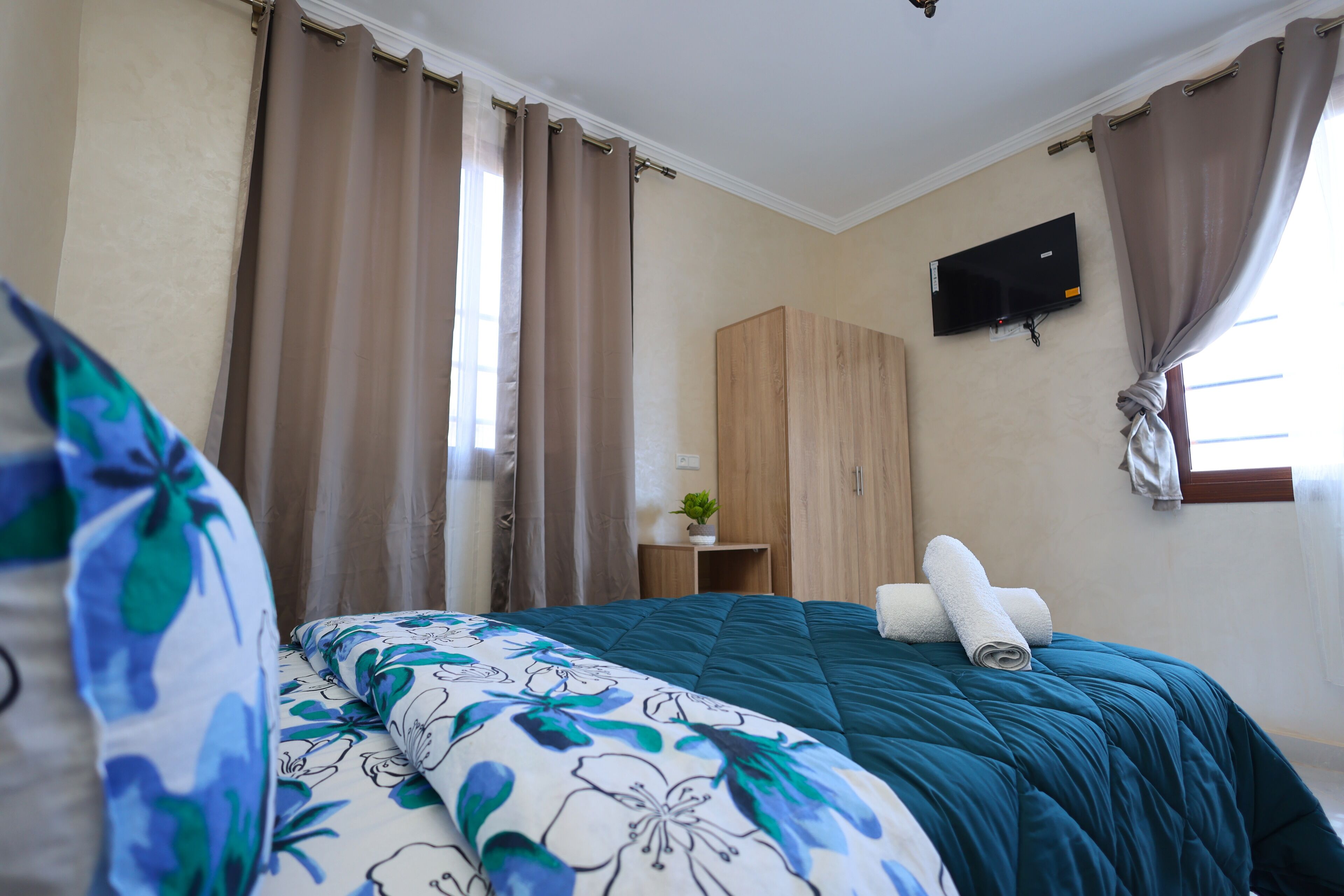 Un dormitorio con cama, cortinas, un televisor montado en la pared y una mesita con una planta.