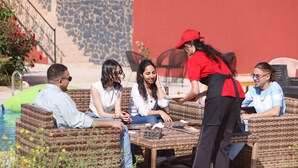 A group of people seated at wicker tables outdoors, with a pool and stone wall in the background. - Hotel palais d'orient (Tafoughalt)