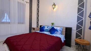Une chambre avec un couvre-lit rouge, une table de chevet avec une lampe, un luminaire fixé au mur et un petit tabouret en bois.