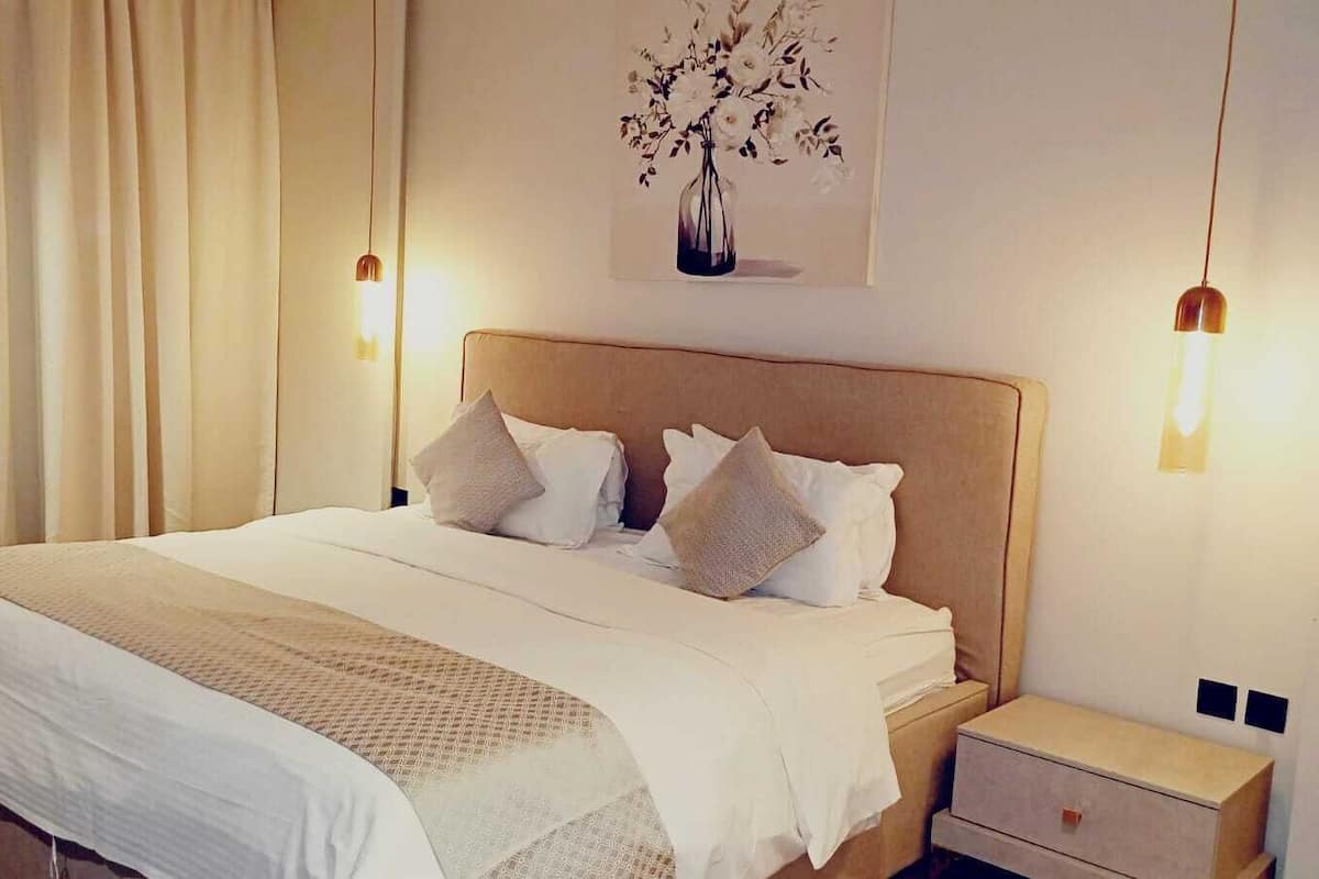 Un lit bien fait, avec du linge de lit blanc, une tête de lit en bois et une table de chevet surmontée d’une lampe.