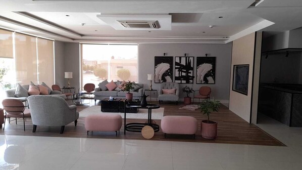 A spacious living room with a grey sofa, pink ottomans, and a coffee table. There are three large abstract paintings on the wall and a fireplace on the right. - ALWURUD ALARBA FOR SERVICED APARTMENTS (Riyadh)