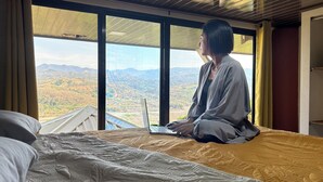 Une personne assise sur un lit, un ordinateur portable sur les genoux, et qui contemple un paysage montagneux à travers de grandes fenêtres.