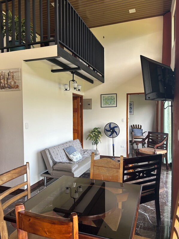 A living room with a glass-top table, wooden chairs, a sofa, a ceiling fan, and a TV mounted on the wall. - Vista Verde (Espíritu Santo)