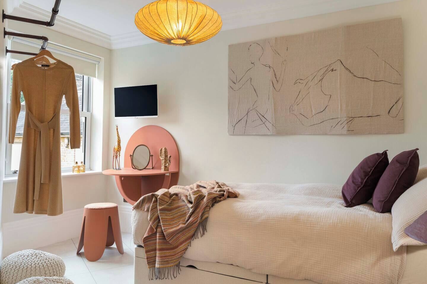 A bedroom with a bed, a hanging robe, a round vanity, a mounted TV, and a large abstract painting.