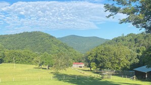 A view from a balcony with a wooden railing, overlooking a green field, a red barn, and a forested mountain range. - Charming 1-bedroom studio in the mountains (Greeneville)