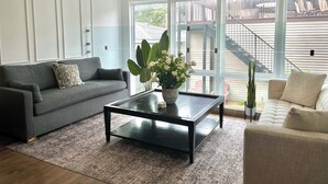 A modern living room with a grey sofa, a black coffee table, and a white armchair. There is a large window with a view of a staircase and outdoor furniture. - 6 BDRM/ 6 BATH LUX Avondale Home, Sleeps 15, Rooftop Deck & 2 Car Garage! (Chicago)