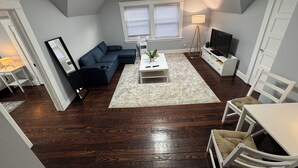 A compact living room with a dark wood floor, a sectional sofa, a white coffee table, a TV stand, and a lamp. - Spacious Yale 2BR with Full Kitchen & WiFi (New Haven)
