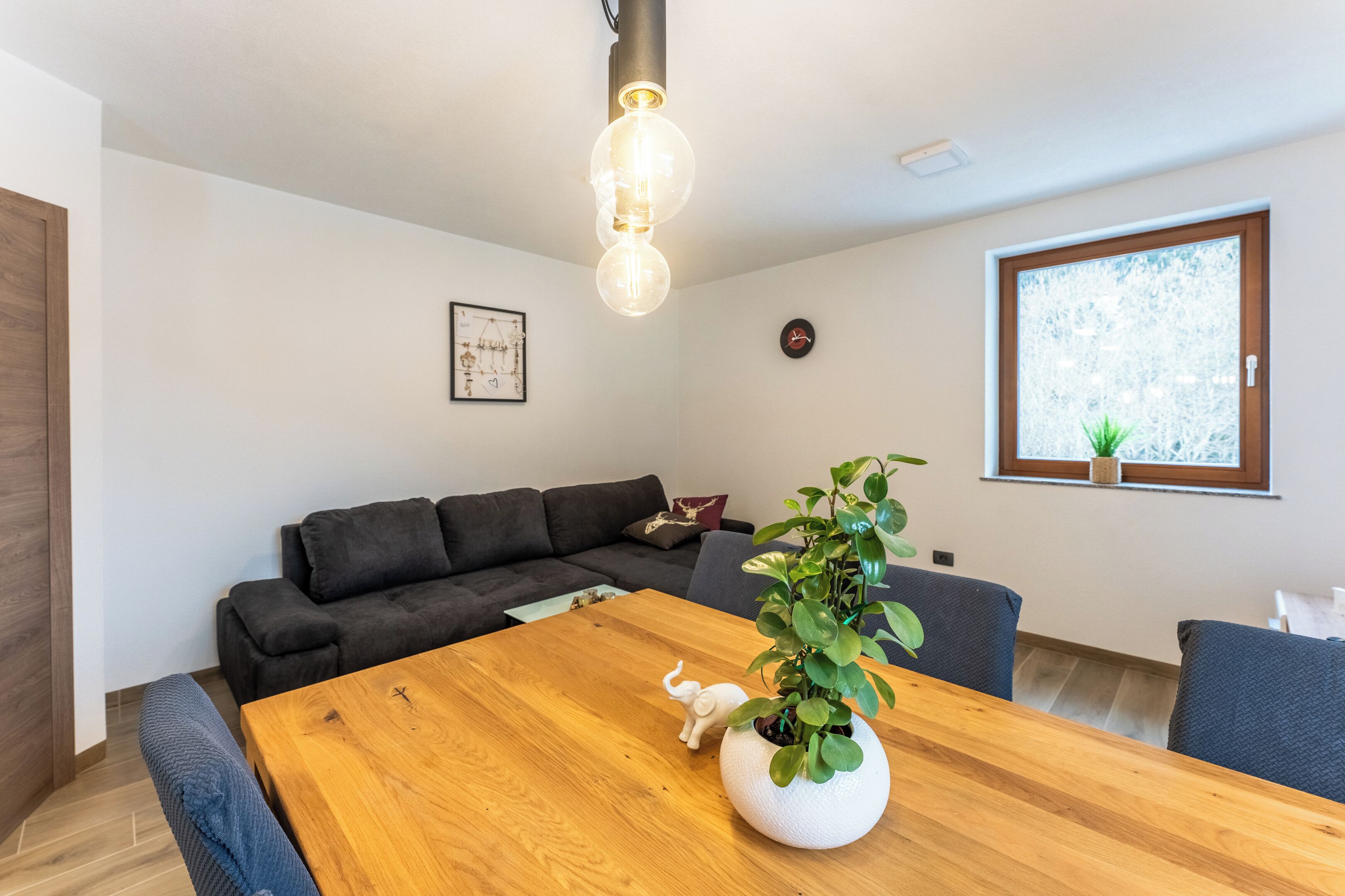 A modern living room with a wooden dining table, dark sofas, a potted plant, and a large window with a view.
