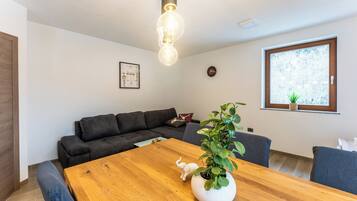 A modern living room with a wooden dining table, dark sofas, a potted plant, and a large window with a view.