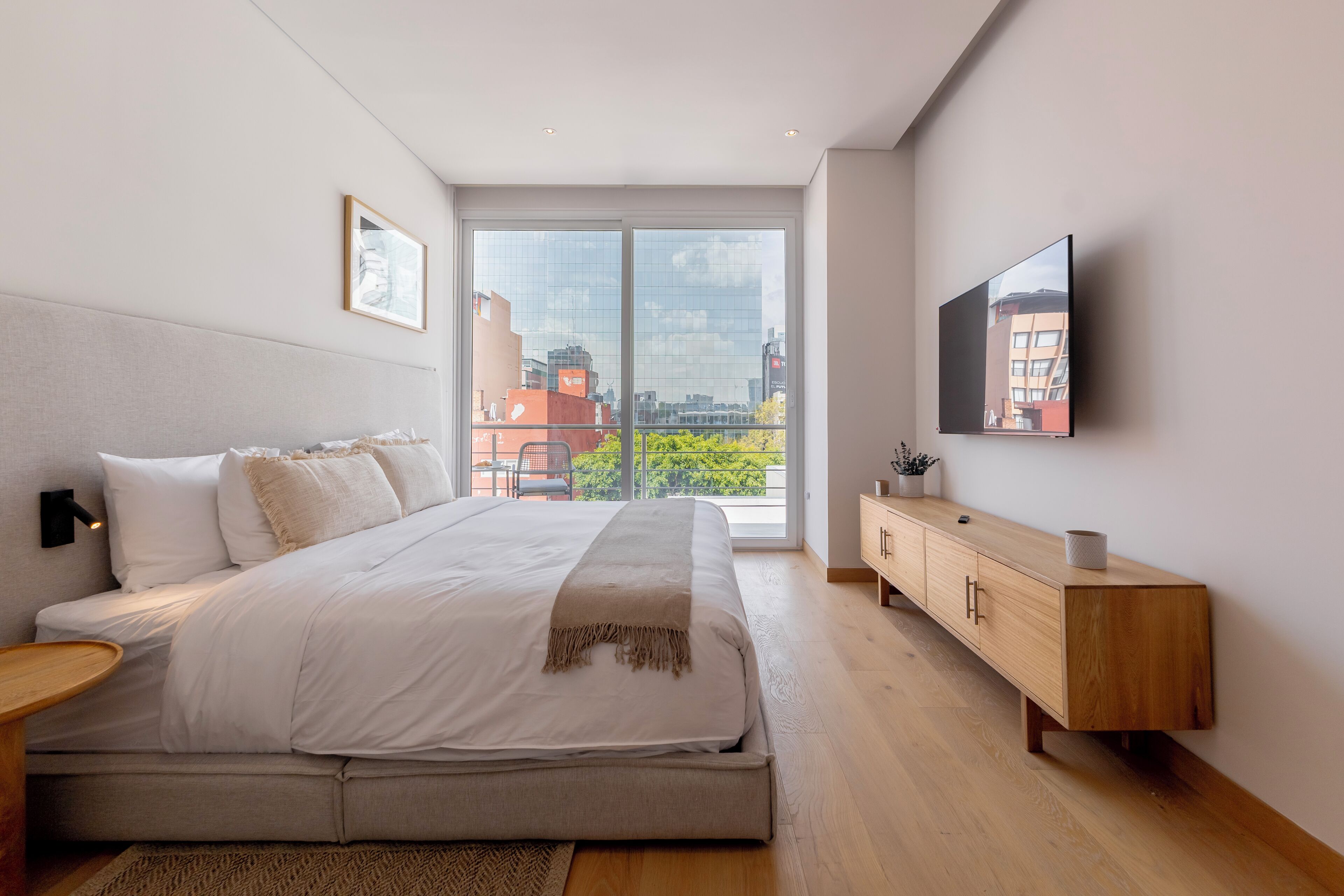 A modern bedroom with a large bed, a wooden nightstand, a TV mounted on the wall, and a view of the city through large windows.
