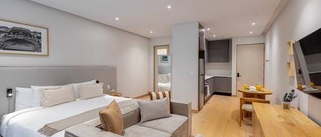 A modern hotel room with a large bed, a sofa, a wooden dining table, and a kitchen area.