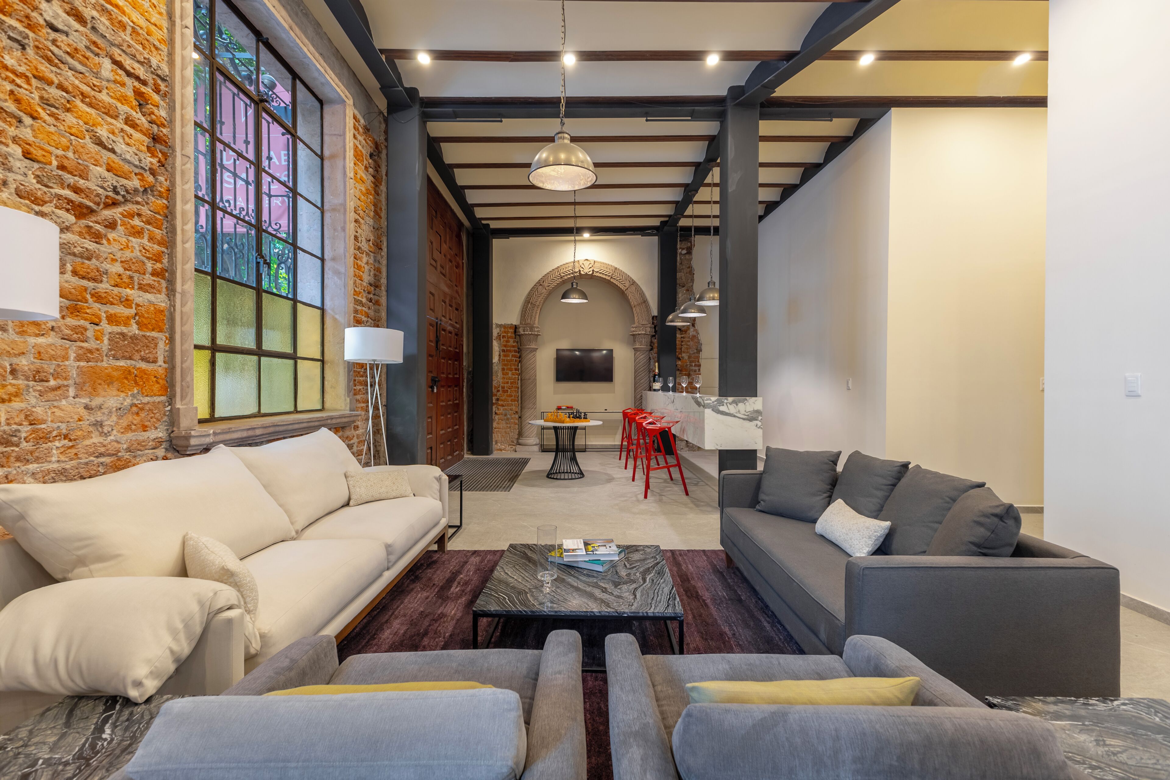 Un salon moderne avec un mur en briques, une grande fenêtre et une cheminée. On y trouve deux canapés, une table basse et un coin repas avec des chaises rouges.