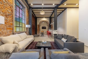 A modern living room with a brick wall, a large window, and a fireplace. There are two sofas, a coffee table, and a dining area with red chairs.