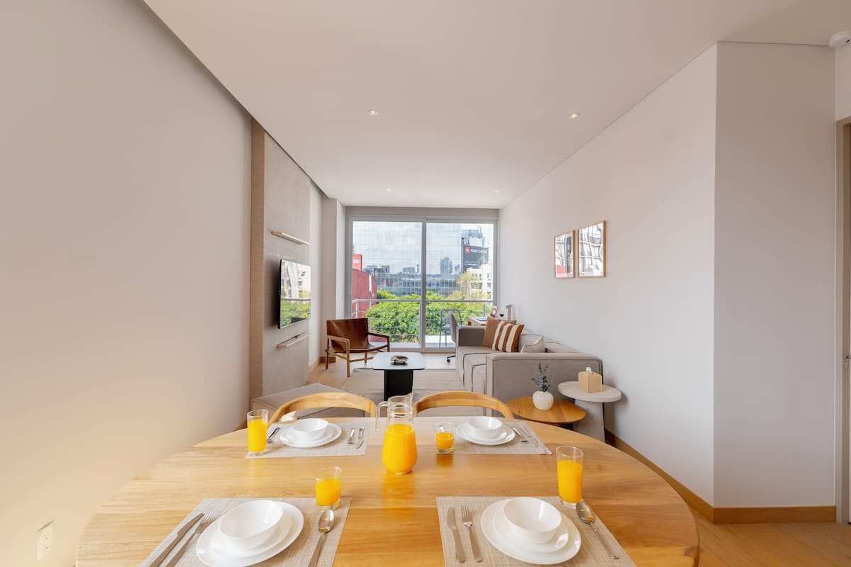 Un comedor con mesa de madera dispuesta para el desayuno, vistas a la ciudad a través de amplios ventanales y una zona de estar acogedora con sofá y sillones.