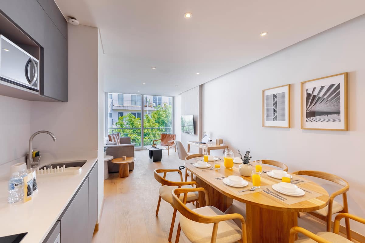 Una cocina moderna con horno empotrado, un comedor con mesa de madera dispuesta para seis personas y amplias ventanas que ofrecen vistas a zonas verdes y edificios.