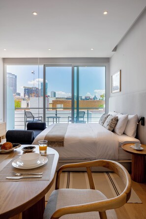 Habitación de hotel con cama, mesa de desayuno con alimentos, una silla y vista a la ciudad a través de amplias ventanas.