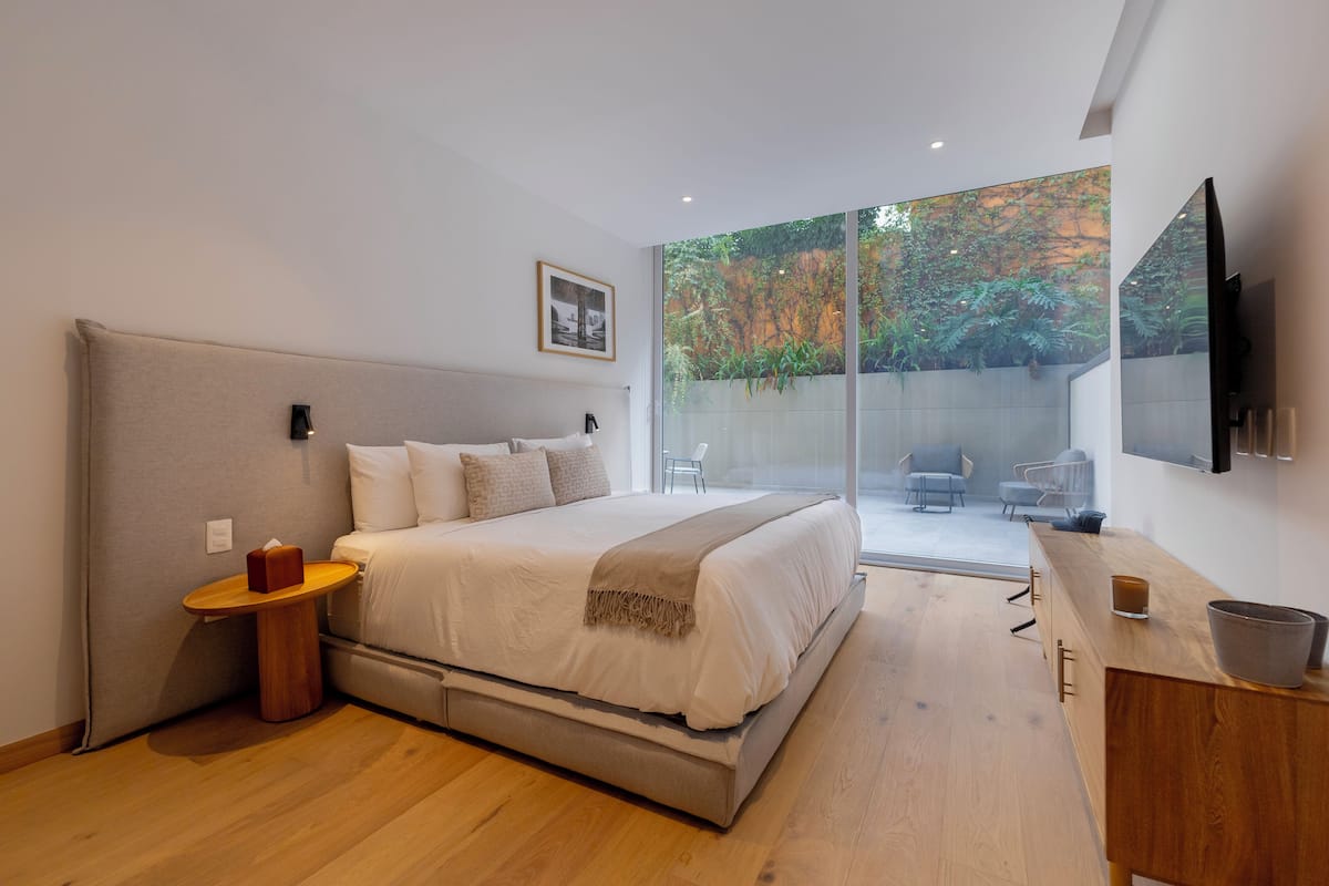 Un dormitorio moderno con una cama grande, una mesita de noche, un aparador de madera y una puerta corrediza de vidrio que da a un espacio exterior con sillas.
