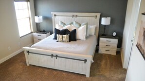 Une chambre à coucher avec un grand lit, deux tables de chevet, une commode et une fenêtre avec des stores.