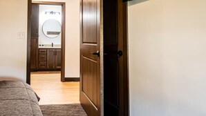 A bedroom with a bed, a wooden door, and a view into a bathroom with a mirror and sink. - Brillion 2‑Bedroom Suite w/ Full Kitchen Near Ariens – Unit #1 (Brillion)