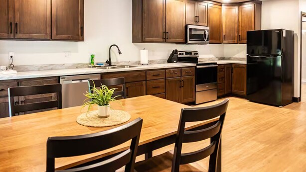 A modern kitchen with wooden cabinets, stainless steel appliances, and a dining table with chairs. - Brillion 2‑Bedroom Suite w/ Full Kitchen Near Ariens – Unit #1 (Brillion)