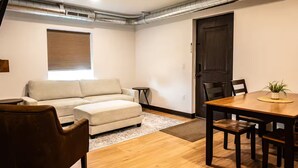 A cozy living room with a sofa, a small side table, a wooden dining table with chairs, and a dark door. - Brillion 2‑Bedroom Suite w/ Full Kitchen Near Ariens – Unit #1 (Brillion)
