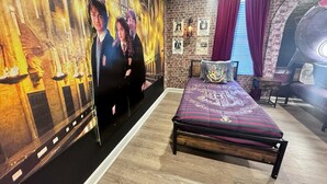A room with a brick wall, a large poster of a wizard and witch, a bed with a Hogwarts crest pillow, and a small table with a lamp.