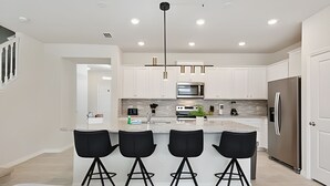 A modern kitchen with white cabinets, a central island with bar stools, stainless steel appliances, and a hanging light fixture.