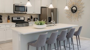 Une cuisine moderne avec des armoires blanches, des appareils en acier inoxydable, un grand îlot central doté de tabourets, un éclairage suspendu et un miroir décoratif en forme de soleil.