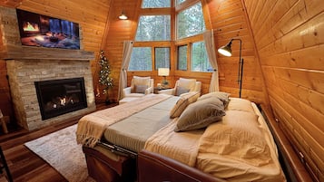 A cozy log cabin bedroom with a fireplace, large windows, and a bed with multiple pillows.
