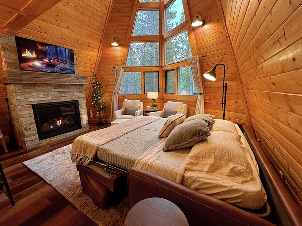 A cozy log cabin bedroom with a fireplace, large windows, and a bed with multiple pillows.