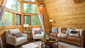 A cozy cabin interior with wooden walls, a brown leather sofa, two armchairs, a coffee table, and large windows offering a view of the forest.