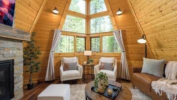 Interior acogedor de una cabaña con chimenea de piedra, paredes de madera y ventanas amplias que dan a un bosque. Hay dos sillones blancos, un sofá marrón y una mesa redonda de centro con una planta y una vela.