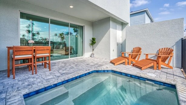 Un espace piscine extérieur moderne, doté d’une porte vitrée donnant sur une terrasse, de chaises en bois et d’une petite table.